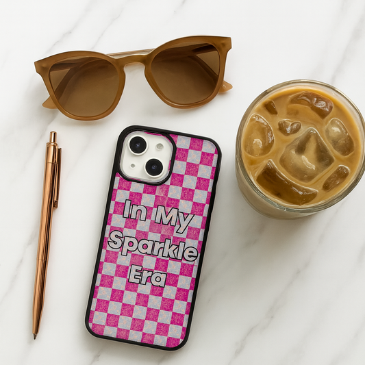 Phone case with 'In My Sparkle Era' text, sunglasses, pen, and iced coffee on a white surface